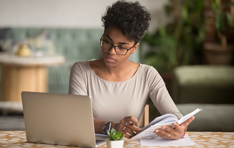 Focused young african american businesswoman or student looking at laptop holding book learning, serious black woman working or studying with computer doing research or preparing for exam online