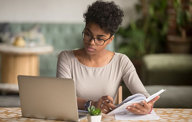 Focused young african american businesswoman or student looking at laptop holding book learning, serious black woman working or studying with computer doing research or preparing for exam online